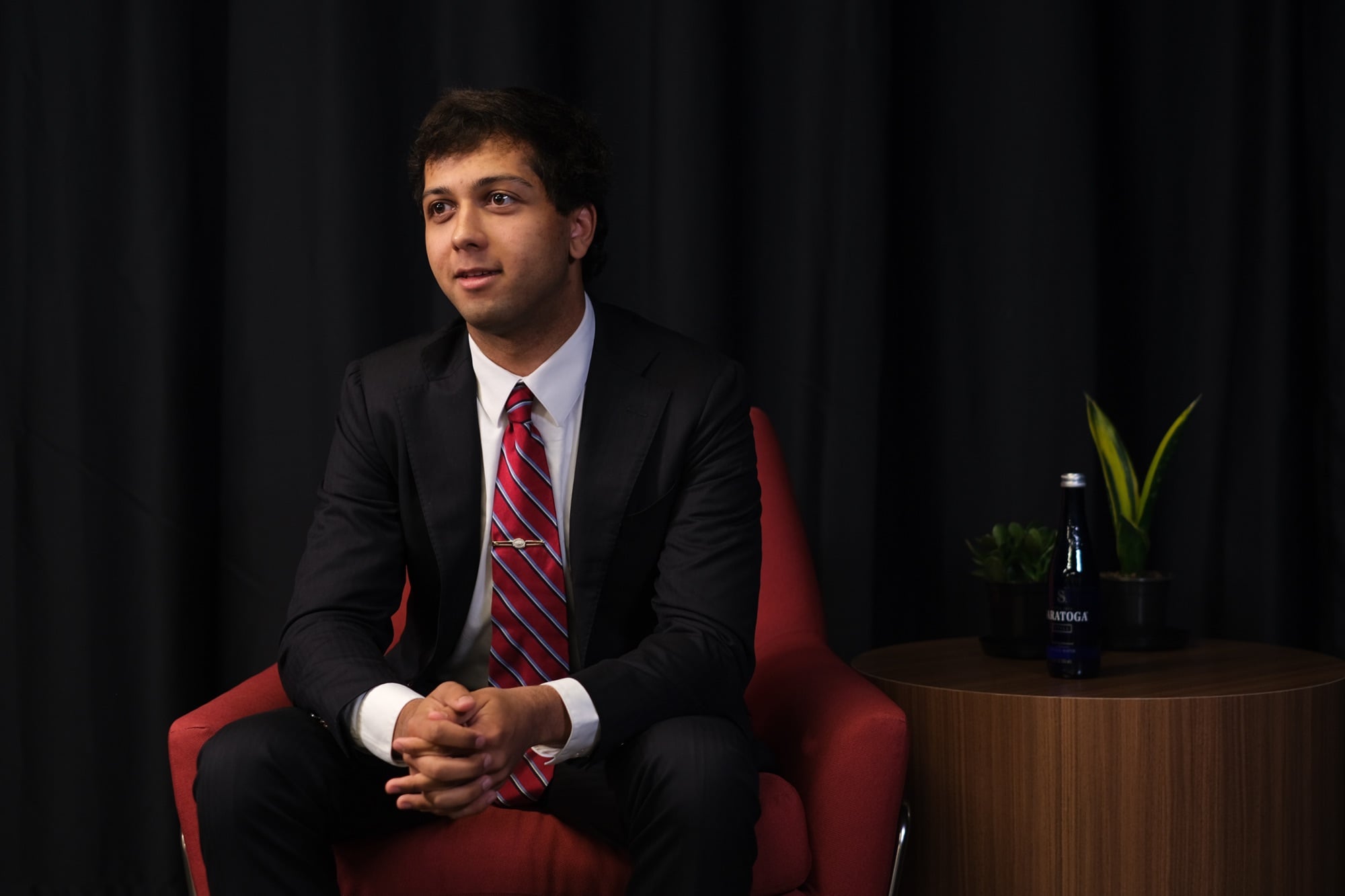 Tejas S. Billa ’28 sits in a red chair against a black curtain.
